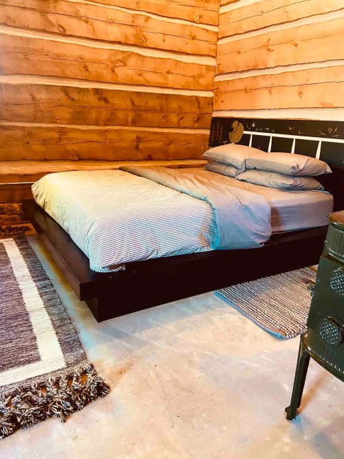 Cozy bedroom with a wooden wall, a simple bed with gray bedding, and a textured rug on the floor.
