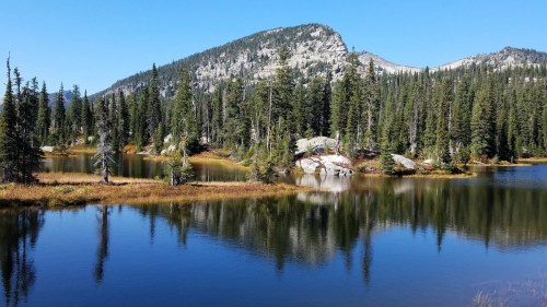 A serene lake surrounded by lush green trees and mountains under a clear blue sky. Reflections shimmer on the water.