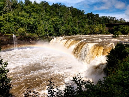 A powerful waterfall cascades over rocks, surrounded by lush greenery and a faint rainbow in the mist.