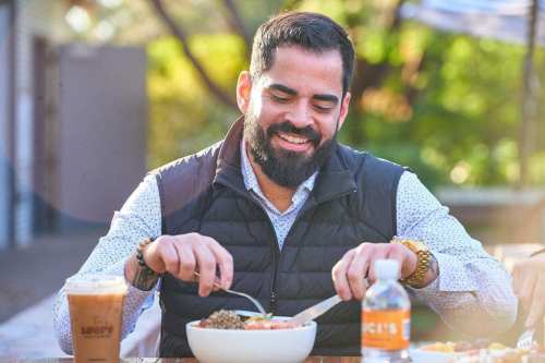 A man with a beard enjoys a meal outdoors, smiling as he eats from a bowl, with a drink beside him.