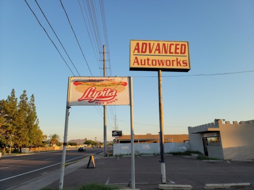 Sign for "Advanced Autoworks" with a banner for "Lupita Hot Dogs" in a clear blue sky setting.