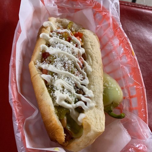 A hot dog in a bun topped with green sauce, diced tomatoes, cheese, and mayonnaise, served in a red and white wrapper.