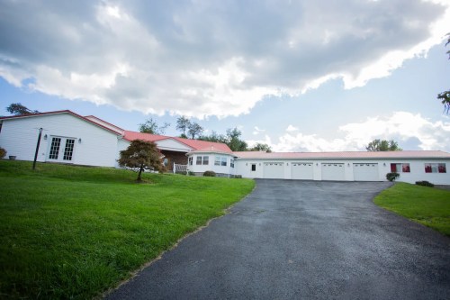 A spacious white house with a red roof, set on a green lawn, with a long driveway leading to a multi-car garage.