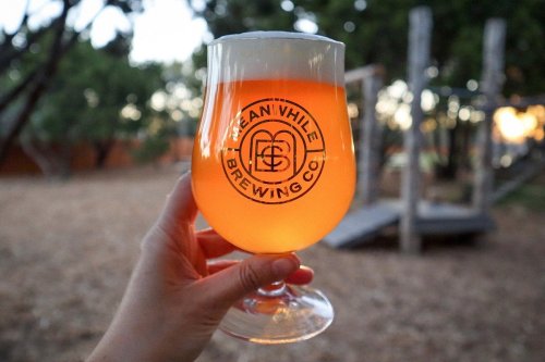 A hand holding a glass of amber beer with a logo, set in a natural outdoor environment.