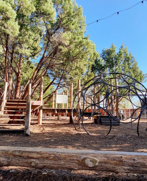 A playground with wooden structures, climbing equipment, and trees under a clear blue sky.