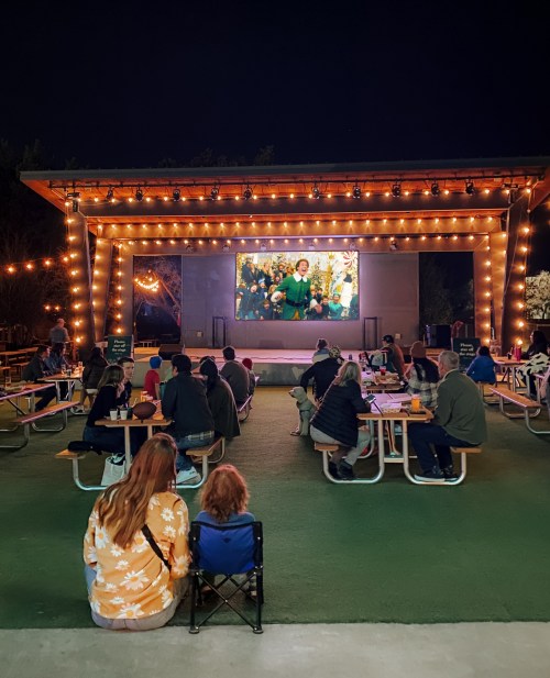 A crowd watches a movie on a large screen outdoors, surrounded by string lights and picnic tables.