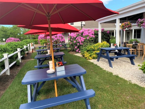 Outdoor dining area with blue picnic tables under red umbrellas, surrounded by blooming flowers and greenery.