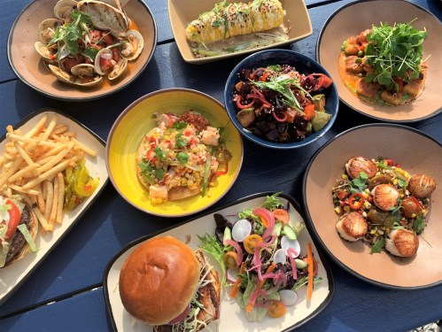 A colorful spread of various dishes including tacos, seafood, salads, fries, and grilled vegetables on blue table.