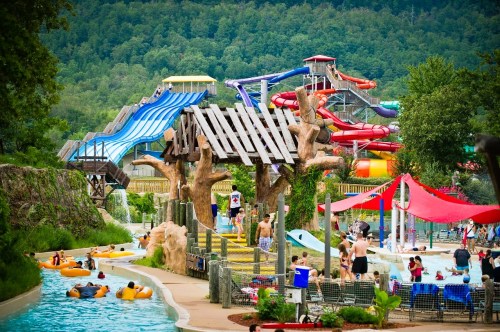 A vibrant water park with slides, lazy river, and families enjoying the attractions amidst lush green hills.