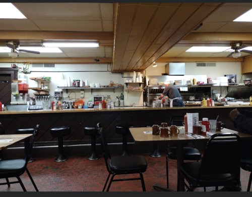 A cozy diner interior with a counter, stools, and a cook preparing food in the kitchen area. Condiments are on the tables.