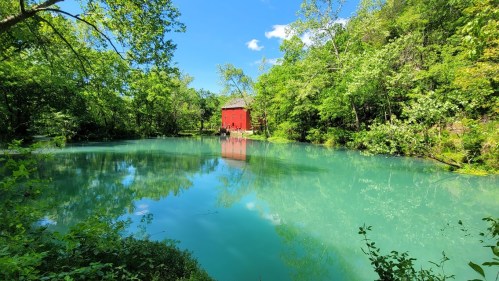 A serene pond surrounded by lush greenery, with a bright red barn reflected in the calm, turquoise water.