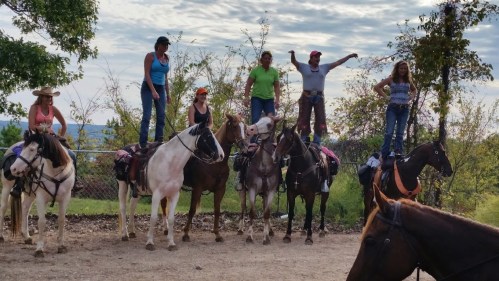 A group of six people standing on horseback, smiling and posing outdoors with trees in the background.