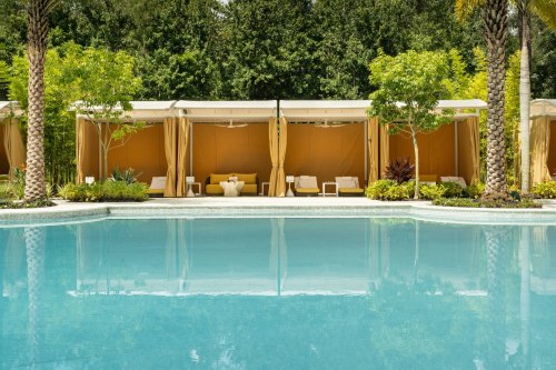A serene pool area with cabanas surrounded by lush greenery and palm trees, reflecting in the clear blue water.