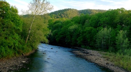 A serene river flows through a lush green landscape, surrounded by trees and rolling hills under a clear sky.