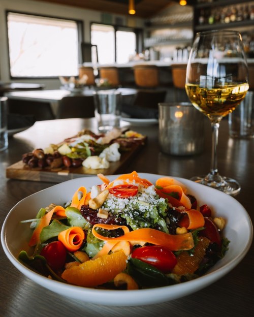A vibrant salad with carrots, tomatoes, and oranges, accompanied by a glass of white wine in a cozy restaurant setting.