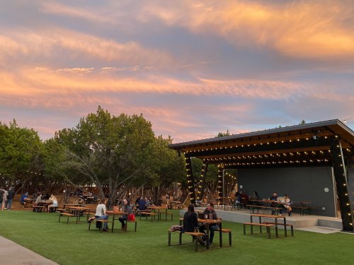 Outdoor venue with picnic tables, string lights, and a colorful sunset sky, surrounded by trees and people enjoying the space.