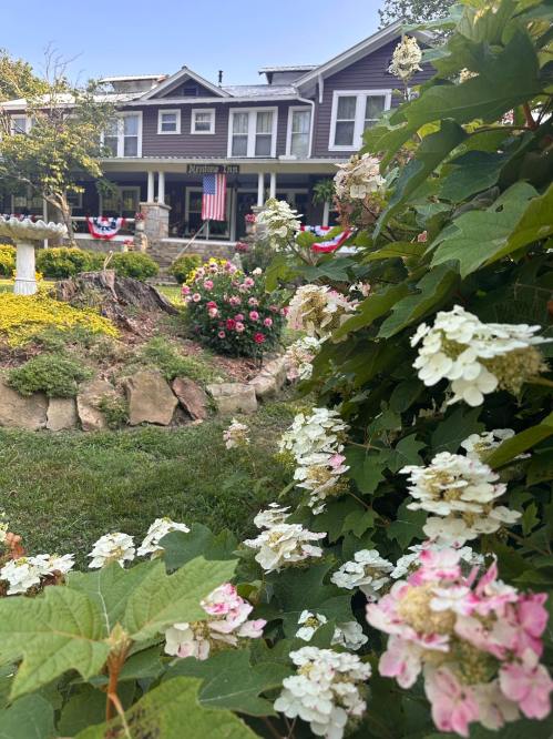 A charming inn with a porch, surrounded by colorful flowers and patriotic decorations, set in a lush garden.