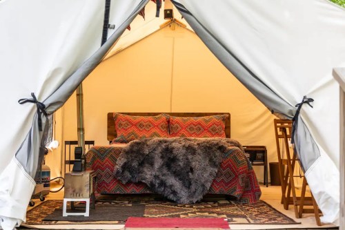 Cozy glamping tent interior featuring a bed with colorful bedding, a faux fur throw, and a small wood stove.