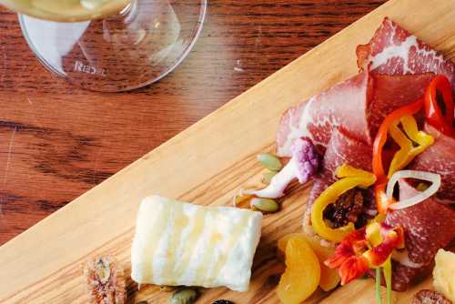 A wooden platter with assorted meats, cheeses, nuts, dried fruits, and colorful peppers, alongside a glass of wine.