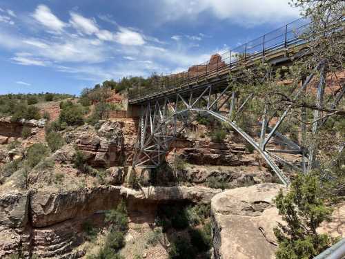 A metal bridge spans a rocky canyon, surrounded by trees and rugged terrain under a blue sky with clouds.