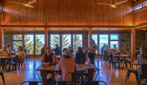 A cozy restaurant interior with wooden walls, large windows, and diners enjoying meals with a scenic view outside.