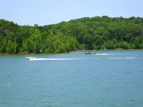 Two boats speed across a calm lake surrounded by lush green trees under a clear blue sky.