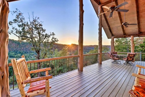 A scenic view from a wooden deck at sunset, overlooking rolling hills and trees. Two rocking chairs are visible.