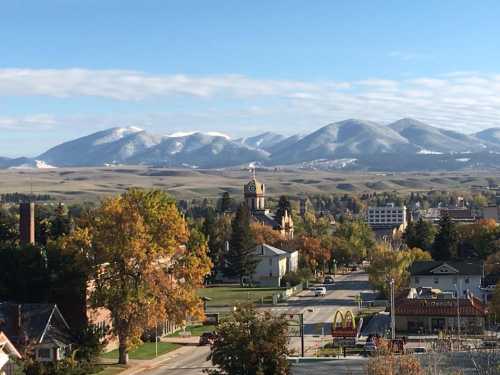 A scenic view of a town with autumn trees, a clock tower, and snow-capped mountains in the background.