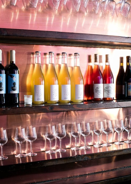 A wooden shelf displays various bottles of wine and sparkling beverages, with elegant wine glasses below.