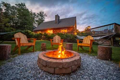 A cozy outdoor fire pit surrounded by wooden chairs, with a log cabin in the background at dusk.