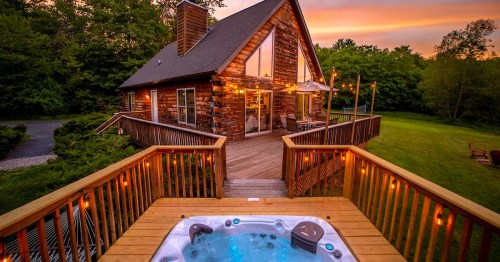 A cozy wooden cabin with a hot tub on the deck, surrounded by greenery and a sunset sky.