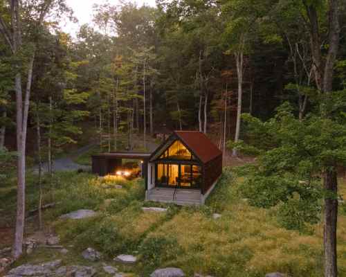 A modern cabin nestled in a forest, surrounded by greenery and rocks, with warm light glowing from its windows at dusk.
