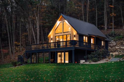 A modern cabin with a sloped roof, large windows, and a deck, surrounded by trees in a serene, autumn setting.