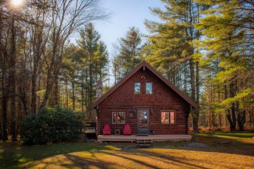 A cozy wooden cabin surrounded by tall trees, featuring a porch with two red chairs in the front.