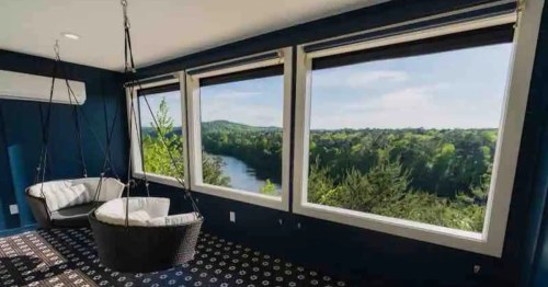 A cozy room with two hanging chairs and large windows showcasing a scenic river view and lush greenery outside.