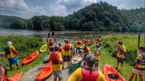 A group of people in life jackets gathers by the river, preparing kayaks for a paddling adventure. Lush greenery surrounds them.