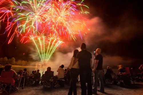A crowd watches colorful fireworks explode in the night sky over a lake, with some people seated in folding chairs.