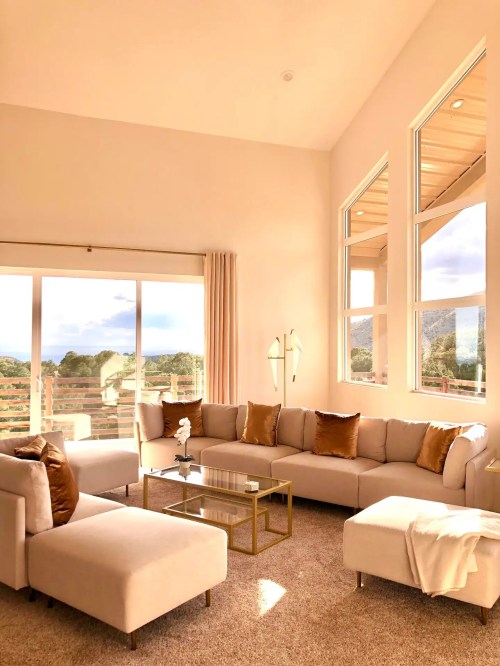 Bright, modern living room with large windows, a beige sectional sofa, gold accents, and a glass coffee table.