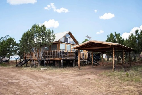 A wooden cabin with a porch and carport, surrounded by trees and a dirt path under a blue sky with clouds.