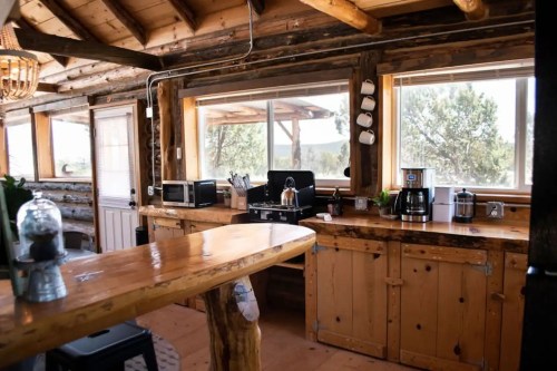 Cozy kitchen with wooden accents, large windows, and appliances like a coffee maker and kettle. Natural light fills the space.