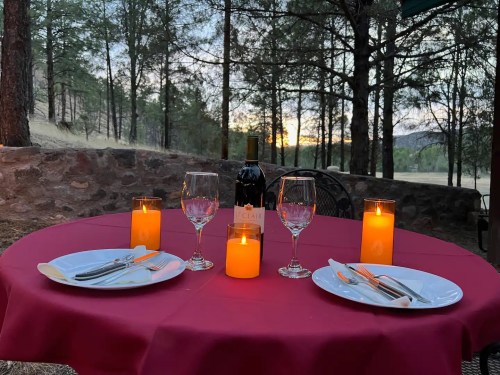 A romantic outdoor dining setup with a red tablecloth, candles, wine, and elegant tableware amidst trees at sunset.