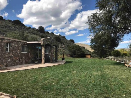 A stone house with a patio overlooks a green lawn and hills under a partly cloudy sky.