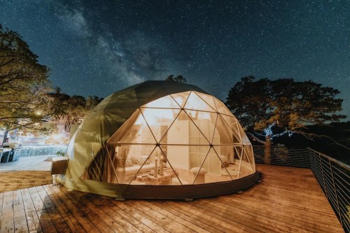 A geodesic dome tent illuminated at night, surrounded by trees under a starry sky.