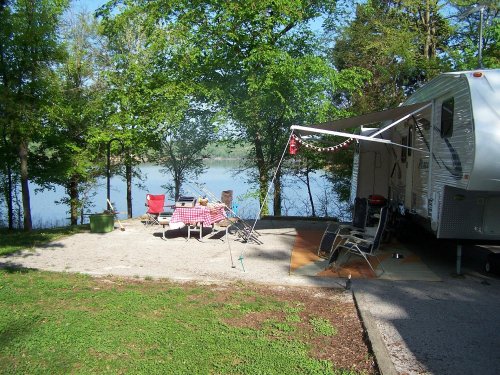 A campsite by a lake featuring a trailer, picnic table, chairs, and trees in a sunny setting.