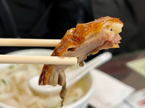 A piece of roasted duck held with chopsticks above a bowl of noodles.