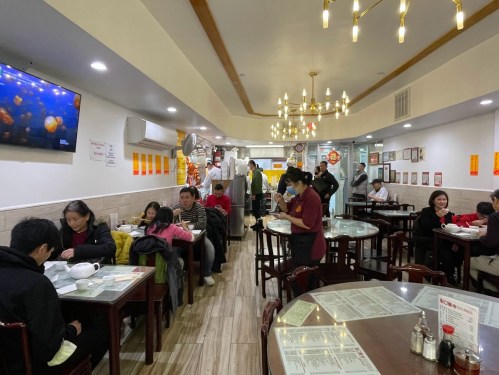 A busy restaurant interior with diners at tables, a TV on the wall, and warm lighting.