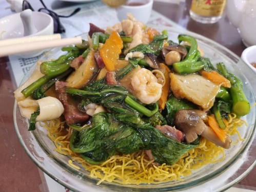 A colorful stir-fried dish with vegetables, seafood, and meat served over crispy noodles in a glass bowl.