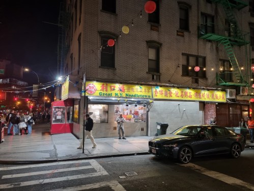 A brightly lit noodle shop at night, with colorful signage and a bustling street scene in the background.