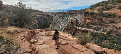 A person walks along a rocky path near a bridge spanning a canyon, surrounded by trees and rugged terrain.