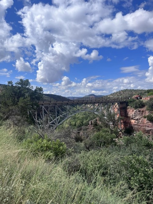 A metal bridge spans a canyon, surrounded by lush greenery and under a blue sky with fluffy clouds.
